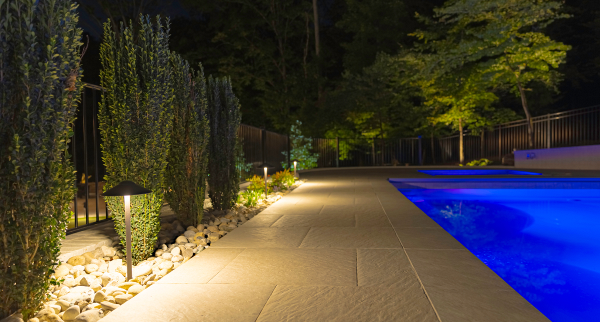 Illuminated poolside walkway beside hedges and a blue-lit pool